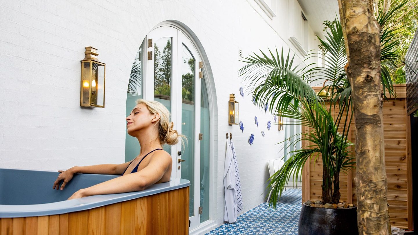 A guest in an outdoor timber tub among blue tiles and palms at Halcyon Wellness
