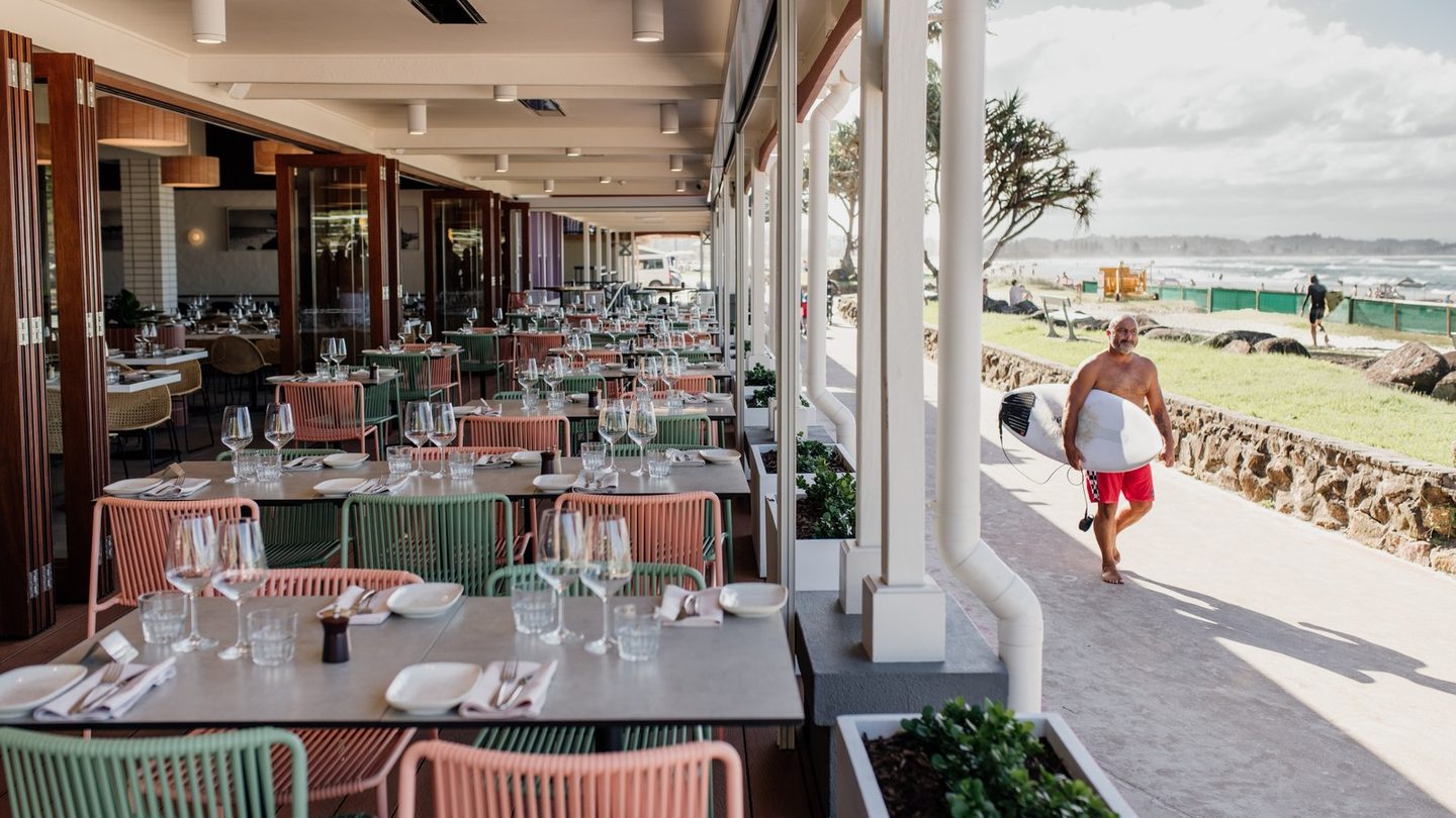 Tables along the beachfront terrace at Siblings Kirra, with a surfer walking past