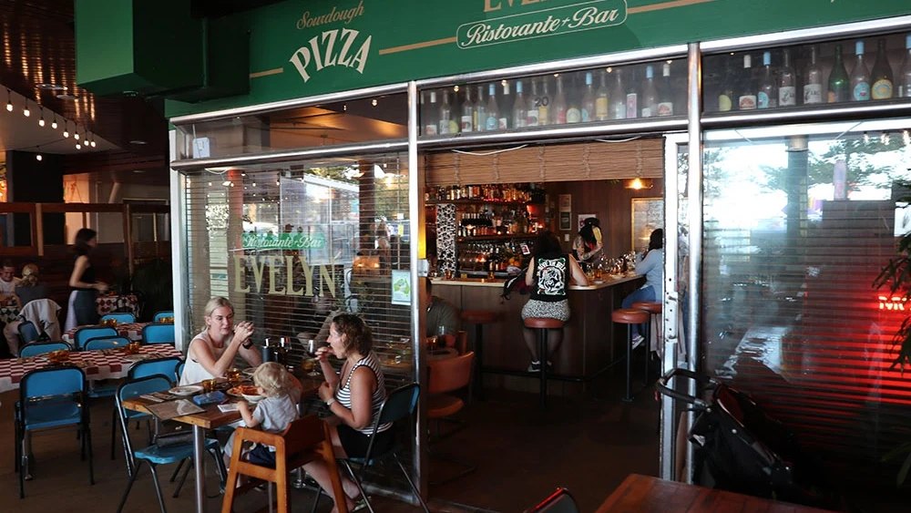 Diners at the open front of Bar Evelyn, under its green pizza sign